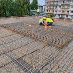 Prace zbrojarskie na dachu budynku wielorodzinnego, widoczna siatka zbrojeniowa z prętów stalowych i pomarańczowe rury, dwóch pracowników w żółtych kamizelkach i kaskach przy pracy, w tle drzewa...