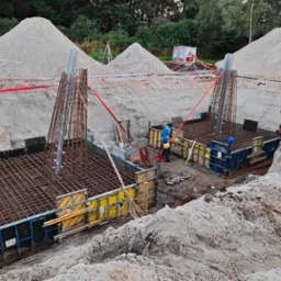 Wykonanie fundamentów pod budynek z użyciem szalunków i zbrojenia, widoczny pracownik w niebieskiej kurtce i koparka Volvo na placu budowy.