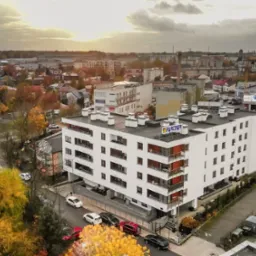Widok z góry na nowoczesny, biały budynek mieszkalny z balkonami i logo 'PARTNER' na dachu, otoczony jesiennymi drzewami i zabudową miejską w promieniach zachodzącego słońca.
