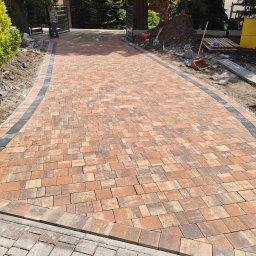 ŁJ--Bód - Nowo ułożona, pomarańczowo-brązowa kostka brukowa z czarnym obramowaniem, prowadząca do bramy wjazdowej, z widocznymi narzędziami i stertą ziemi w tle.