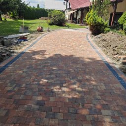 ŁJ--Bód - Nowo ułożona, zakrzywiona ścieżka z kostki brukowej w odcieniach czerwieni i brązu z czarnym obramowaniem, prowadząca do domu z czerwonym dachem. Na trawniku widoczne narzędzia i materiały budowlane.