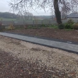 ŁJ--Bód - Świeżo ułożona ścieżka z szarej kostki brukowej z czarnym obrzeżem, prowadząca od bramy wjazdowej z czerwonym traktorem do ogrodu z drewnianym płotem, widok na wiejski krajobraz w pochmurny dzień.