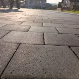 Świeżo ułożona, szara kostka brukowa na podjeździe w słoneczny dzień, widoczne ślady po pracy wykonawców i odległe zabudowania mieszkalne.