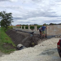 ŁJ--Bód - Budowa ogrodzenia z beżowych bloczków betonowych, widoczni dwaj mężczyźni w trakcie pracy, taczka i narzędzia obok, w tle krajobraz z drzewami i pochmurnym niebem.