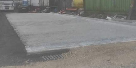 Świeżo wylana, szara, betonowa powierzchnia na zewnątrz, z widoczną kratką ściekową i fragmentami samochodów w tle.