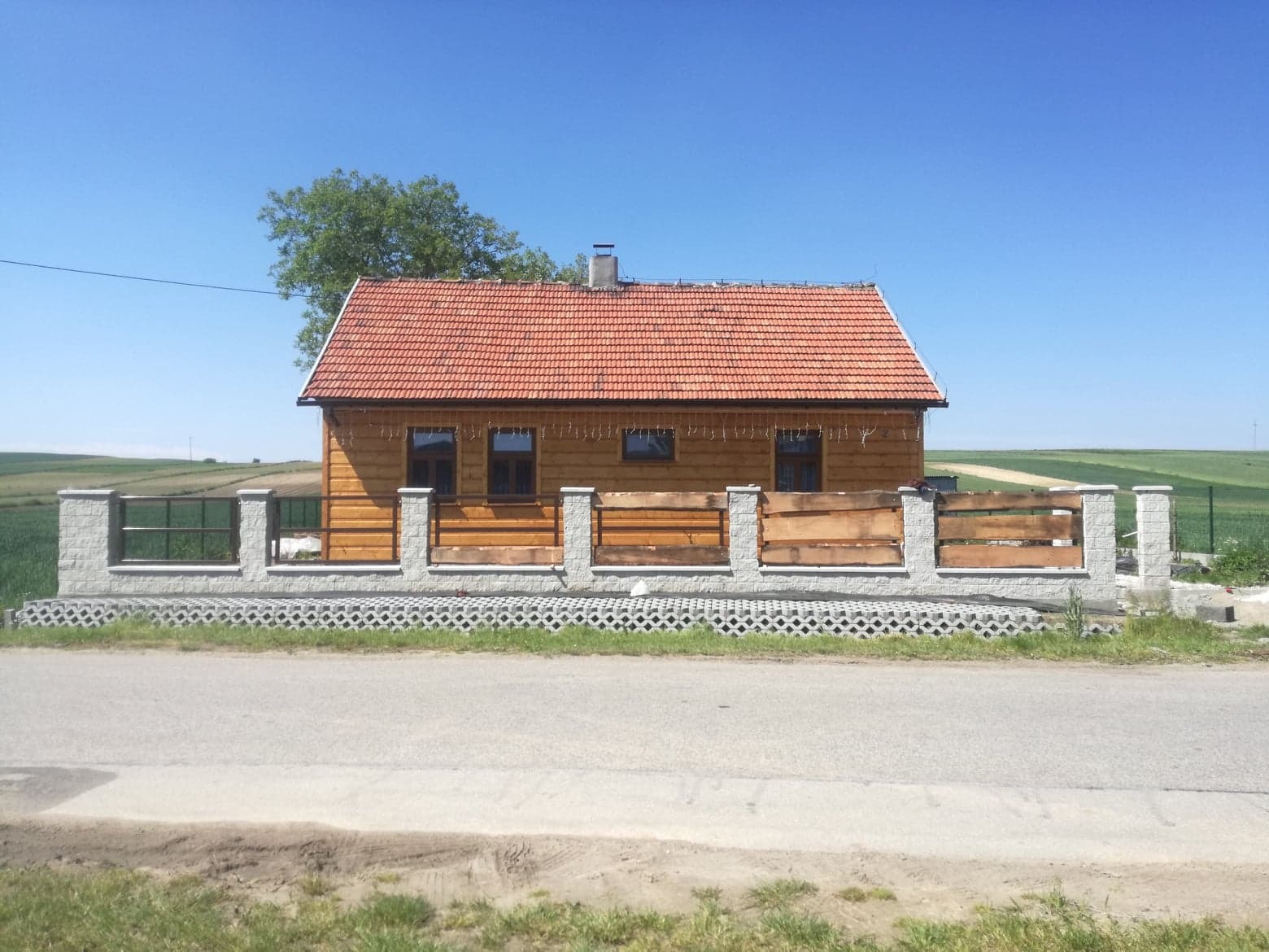 Drewniany dom z czerwonym dachem i ogrodzeniem z drewnianych paneli oraz betonowych słupków, widok z poziomu ulicy w słoneczny dzień.