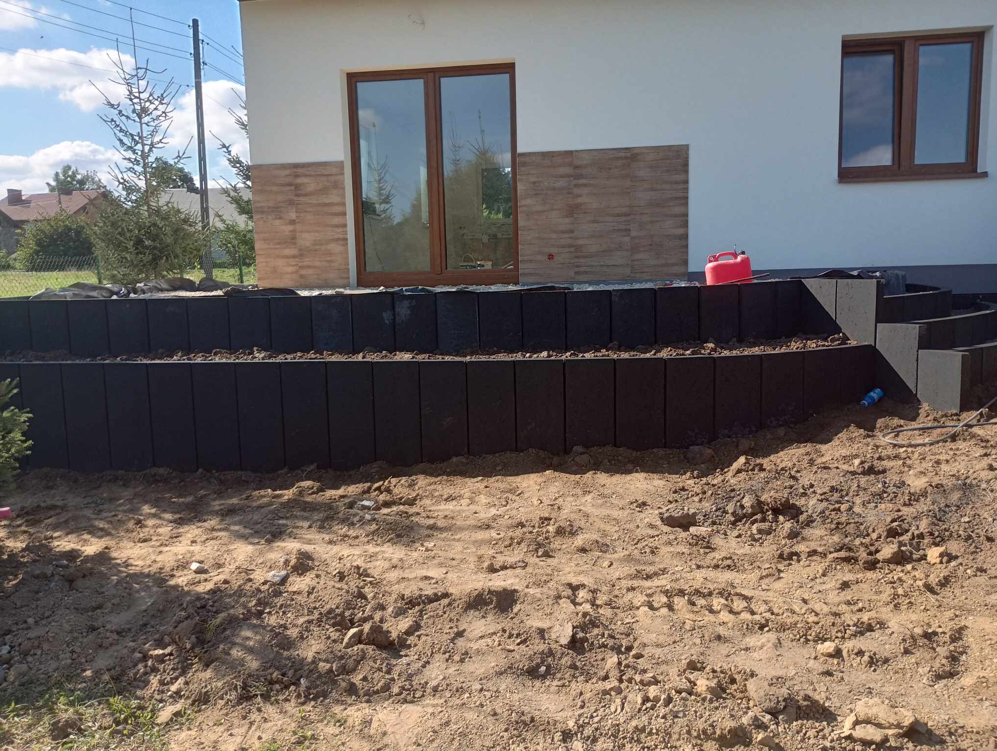 Budowa schodów z ciemnych, betonowych elementów przed jasnym domem z brązowymi oknami i dekoracyjnymi panelami ściennymi, widoczny czerwony kanister i rozsypana ziemia.