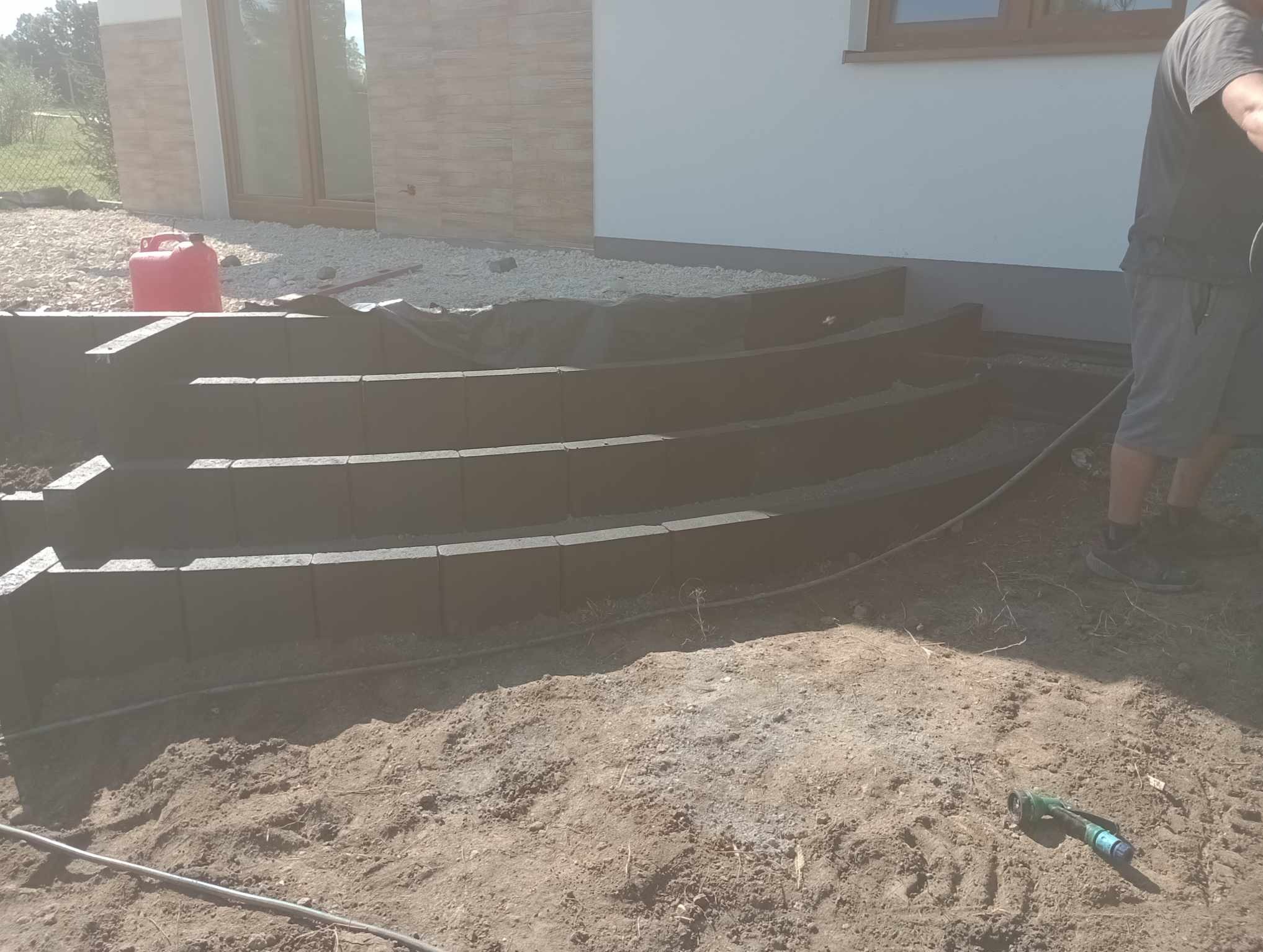 Budowa schodów zewnętrznych z betonowych elementów obrzeżowych, widoczny częściowo pracownik, obok budynku z oknem.