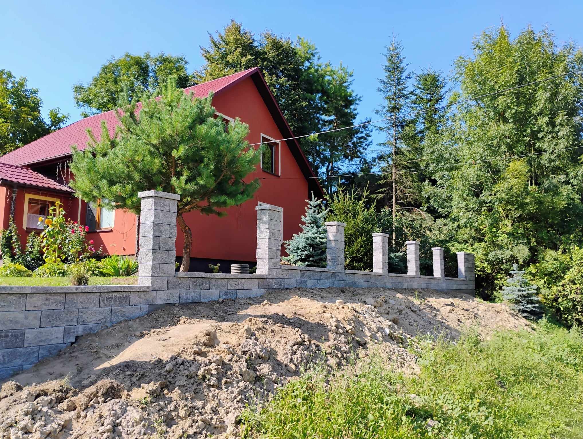 Niski murek z szarej kostki betonowej z filarami, w trakcie budowy, z widoczną ziemią i roślinnością, na tle czerwonego domu i zieleni drzew.