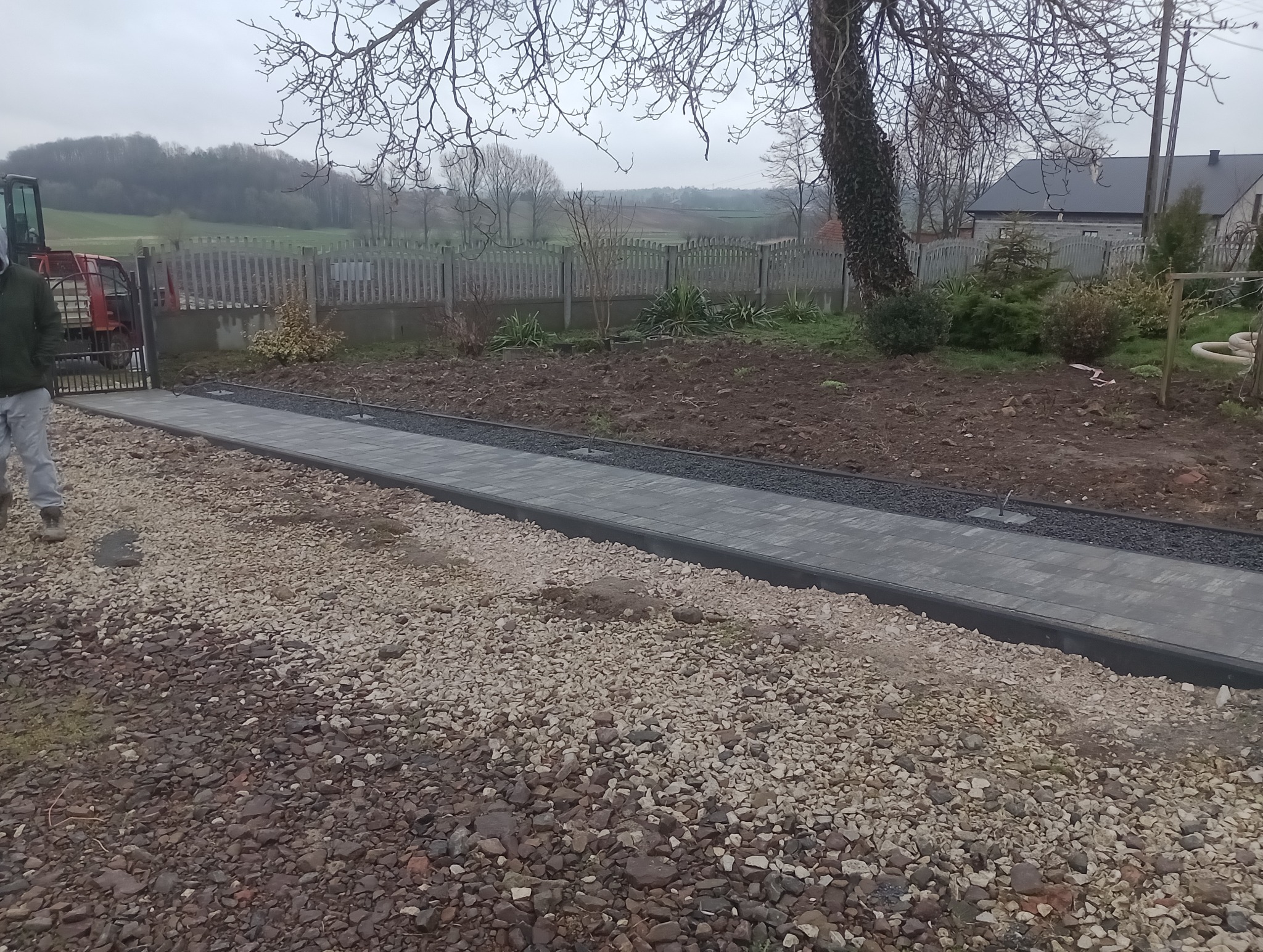 Świeżo ułożona ścieżka z szarej kostki brukowej z czarnym obrzeżem, prowadząca od bramy wjazdowej z czerwonym traktorem do ogrodu z drewnianym płotem, widok na wiejski krajobraz w pochmurny dzień.