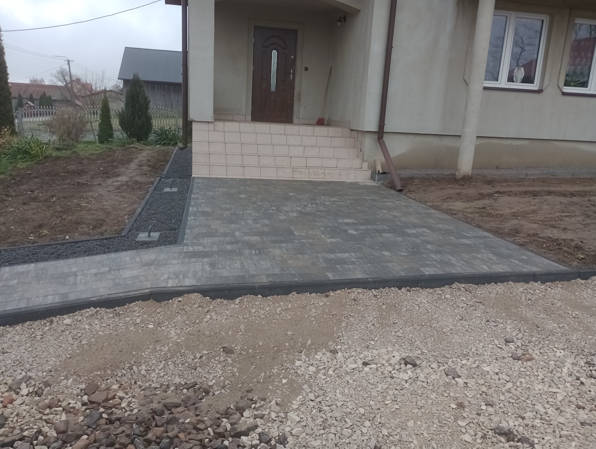 Nowo ułożona, szara kostka brukowa prowadząca do schodów wejściowych domu jednorodzinnego, obrzeża z ciemnego kamienia, fragment rabaty z drobnym, czarnym żwirem.