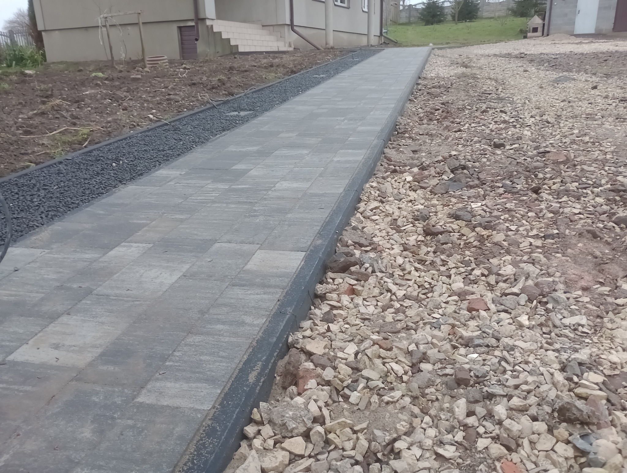 Świeżo ułożony chodnik z szarej kostki brukowej, obrzeża z czarnego grysu, teren wokół jeszcze niezagospodarowany, widoczne kamienie i gruz.