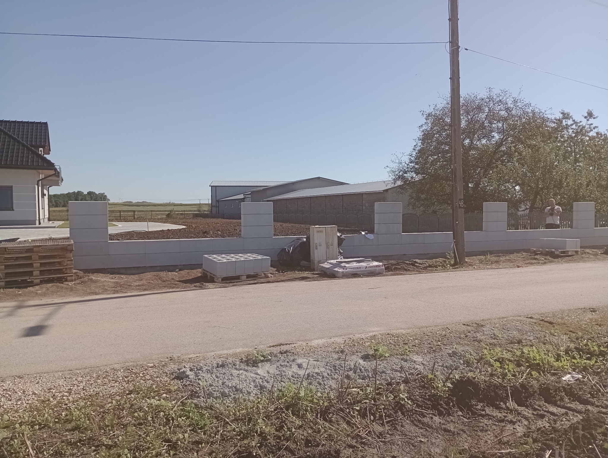 Wznoszenie ogrodzenia z prefabrykatów betonowych: widok na częściowo postawiony mur, palety z bloczkami, zapakowane elementy, narzędzia i pracująca osoba w tle.