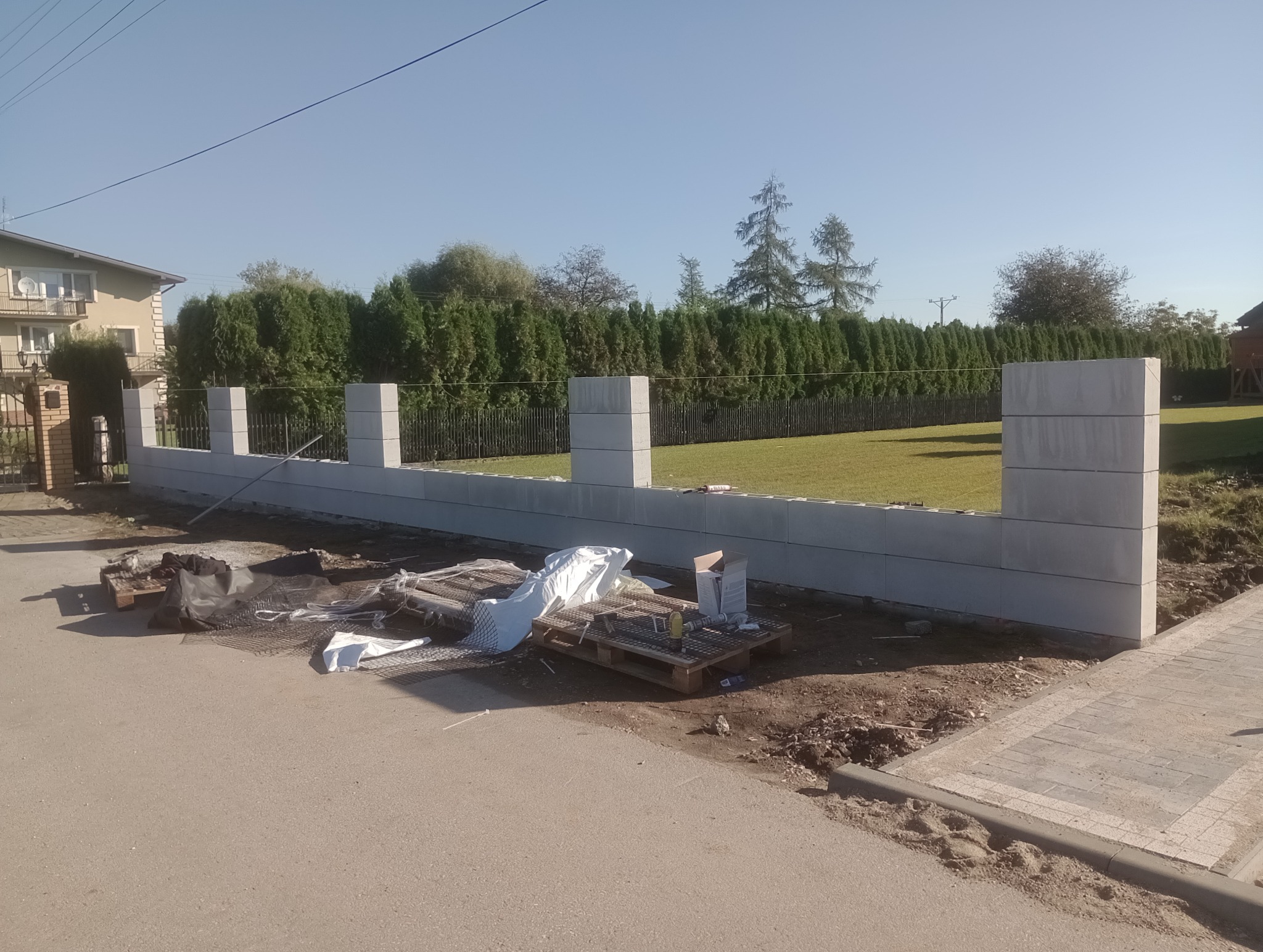 Wznoszony mur z prefabrykowanych betonowych elementów ogrodzeniowych, widoczne palety z materiałami budowlanymi i narzędziami na tle zieleni ogrodu i domu.