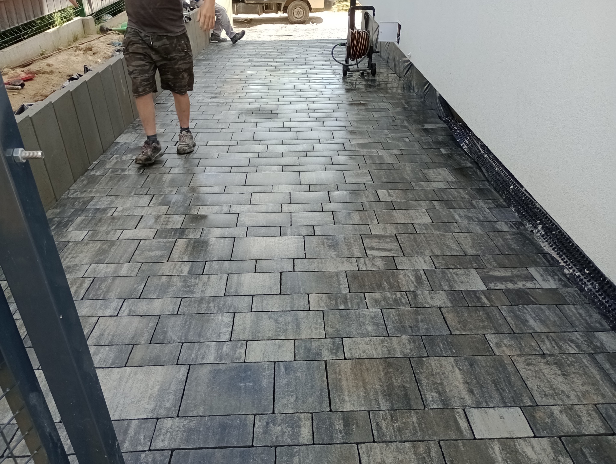 Świeżo ułożona, szara kostka brukowa w kształcie prostokątów, widoczny fragment ogrodzenia oraz pracownik w tle, obok biała ściana budynku.