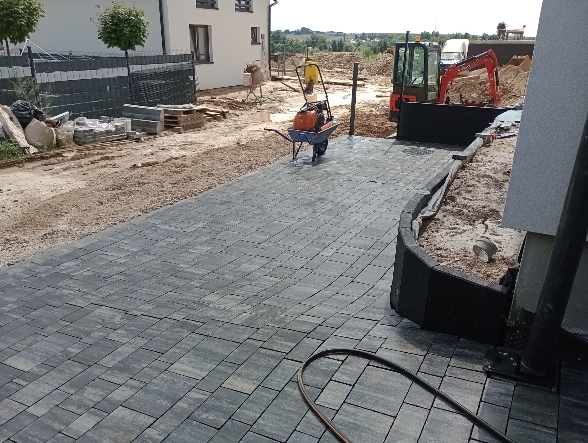 Ułożona szara kostka brukowa na podjeździe, widoczna zagęszczarka i taczka, w tle mini koparka i mieszalnik do betonu, prace w toku.