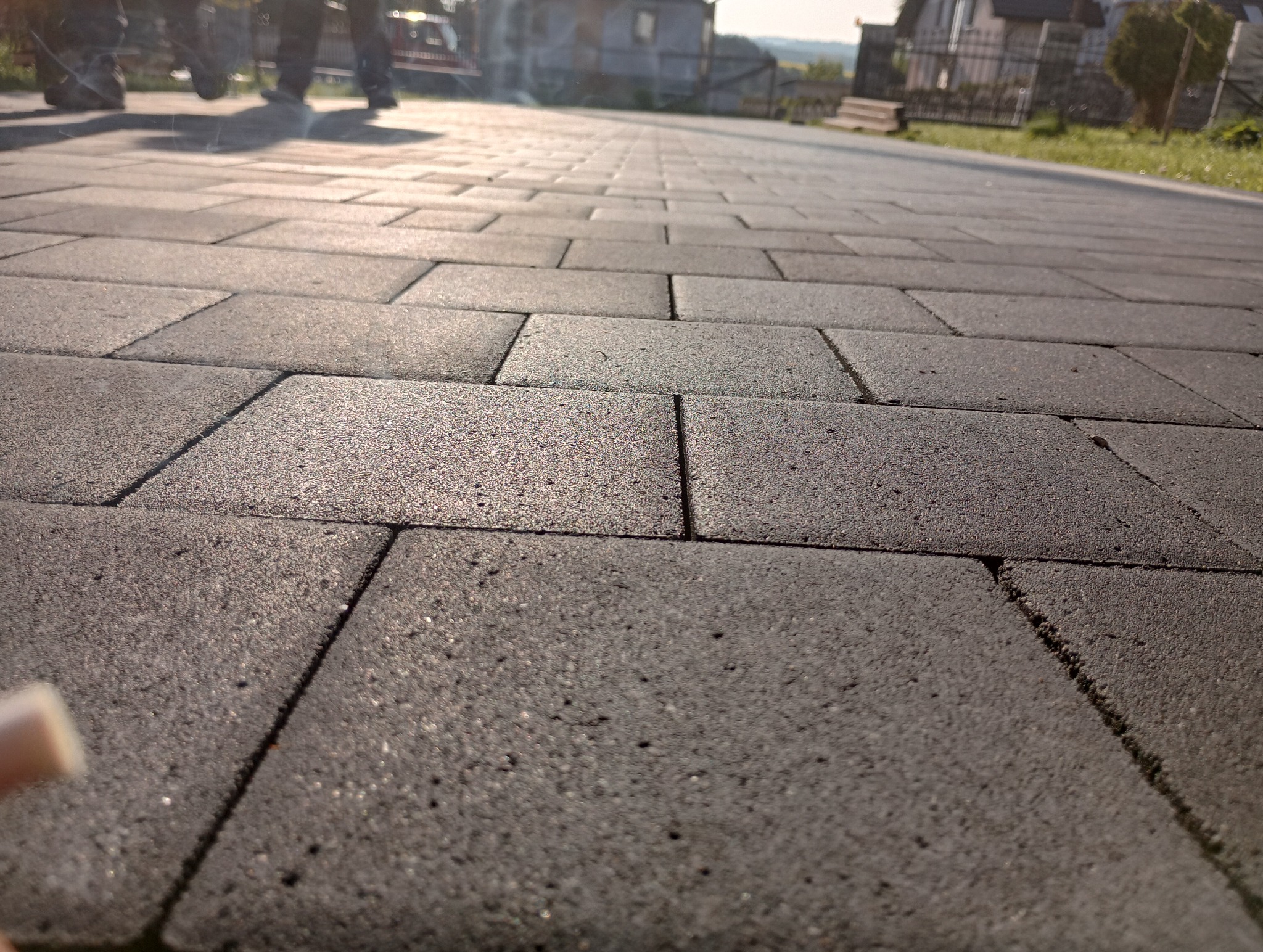 Świeżo ułożona, szara kostka brukowa na podjeździe w słoneczny dzień, widoczne ślady po pracy wykonawców i odległe zabudowania mieszkalne.
