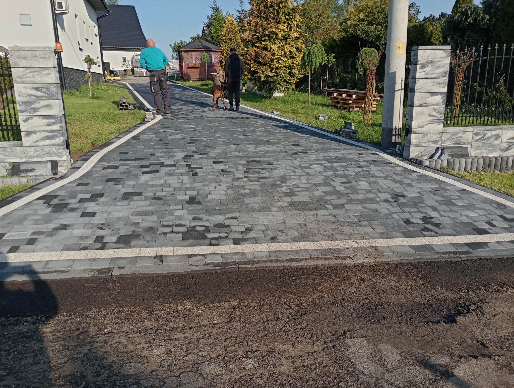 Ułożona kostka brukowa w kolorach szarości i czerni tworząca wzór na podjeździe, obramowana jasnymi kostkami, z widocznym fragmentem asfaltu przed wjazdem i dwoma mężczyznami z psem w tle.