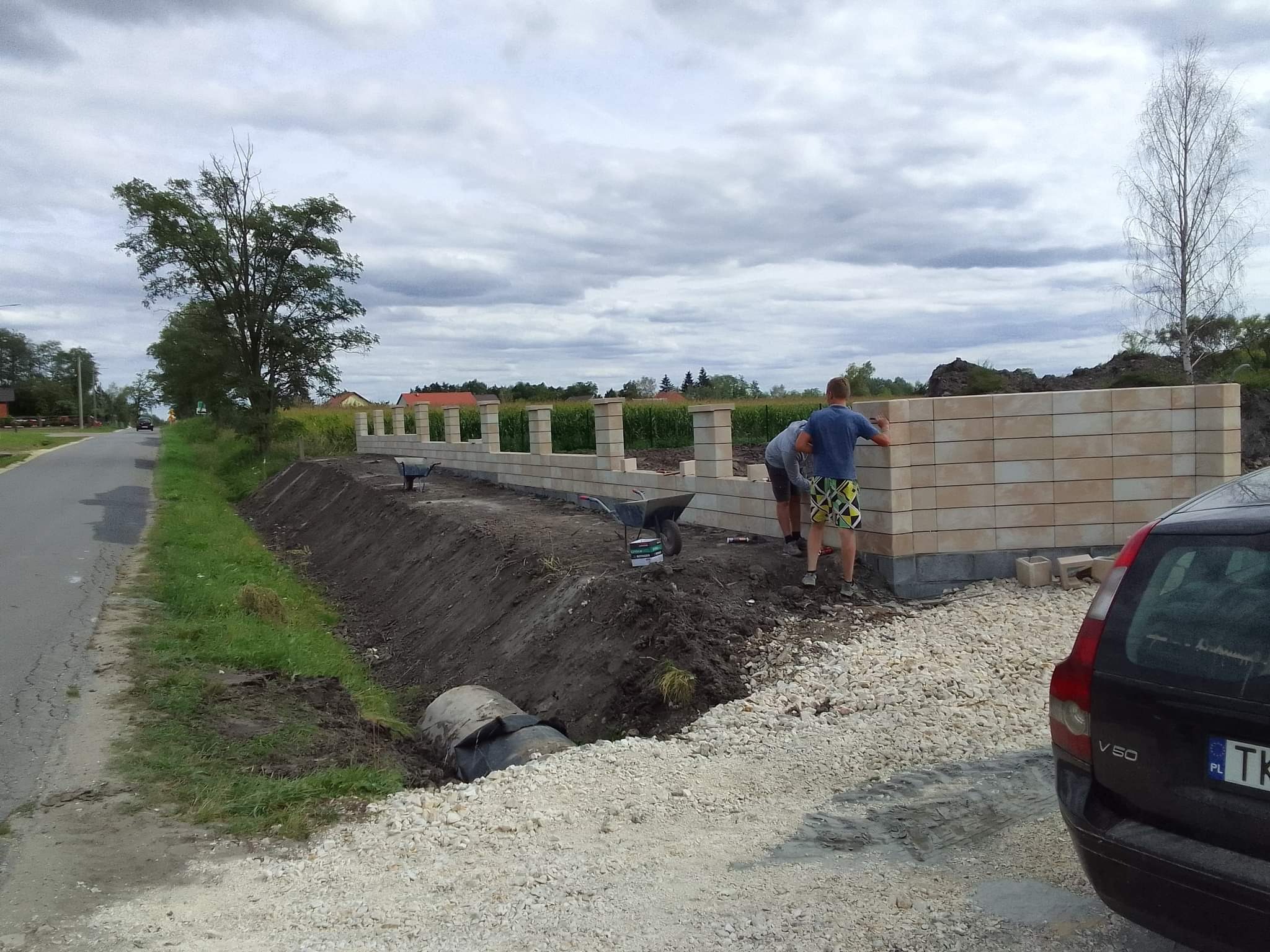 Budowa ogrodzenia z beżowych bloczków betonowych, widoczni dwaj mężczyźni w trakcie pracy, taczka i narzędzia obok, w tle krajobraz z drzewami i pochmurnym niebem.