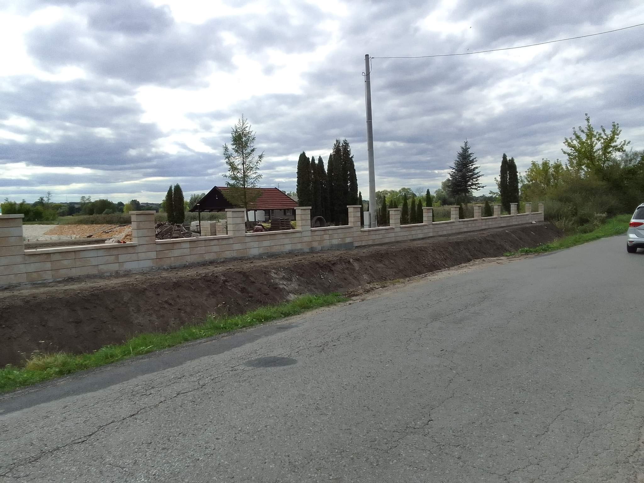 Długi, niski murek z beżowych bloczków betonowych z filarami, oddzielający posesję od drogi; w tle widoczne tuje i dom z czerwonym dachem.