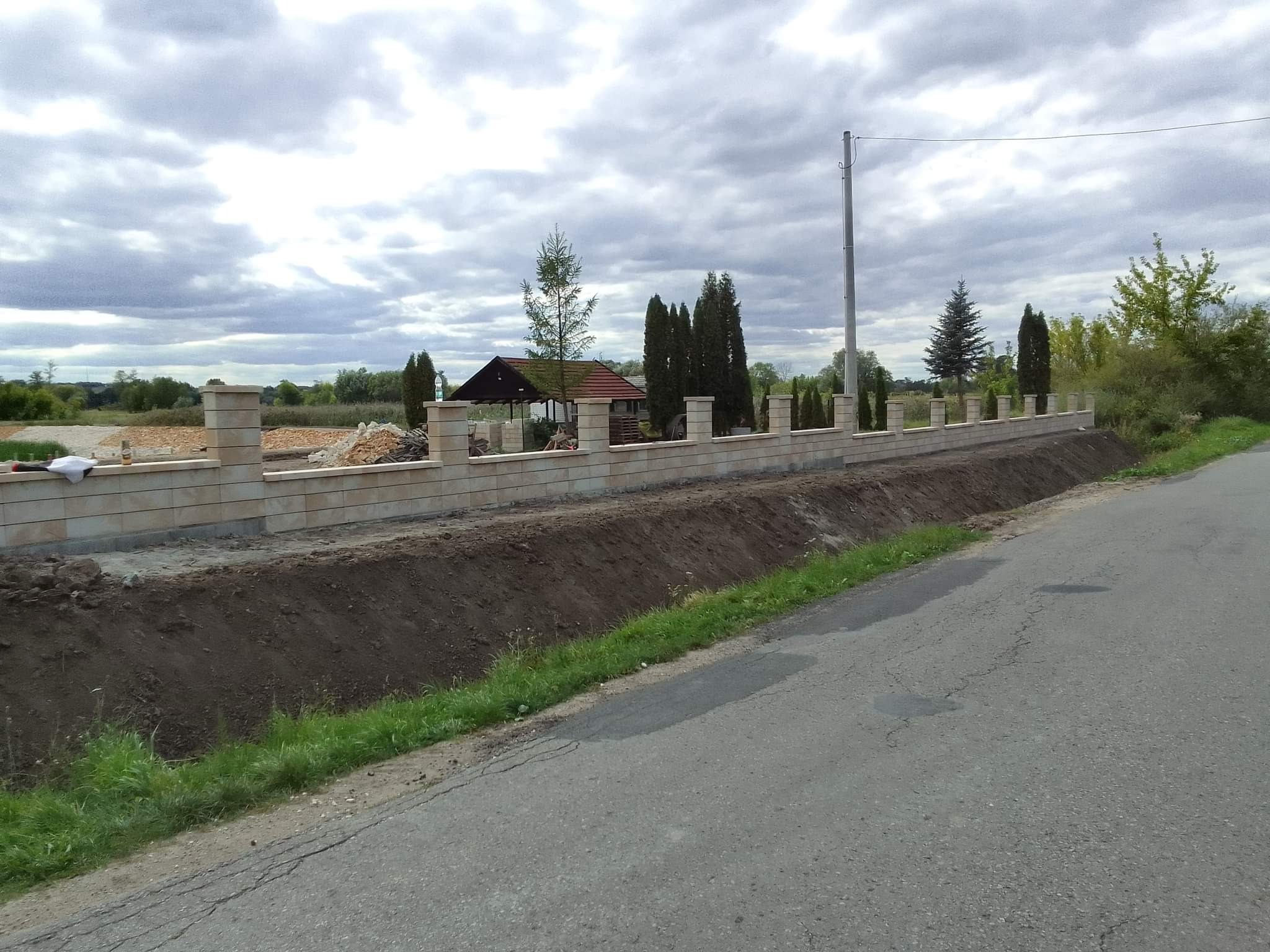 Wznoszony niski murek z prefabrykatów betonowych z kolumnami, widok z boku drogi, przygotowany teren pod dalszą budowę, w tle budynek gospodarczy i drzewa.