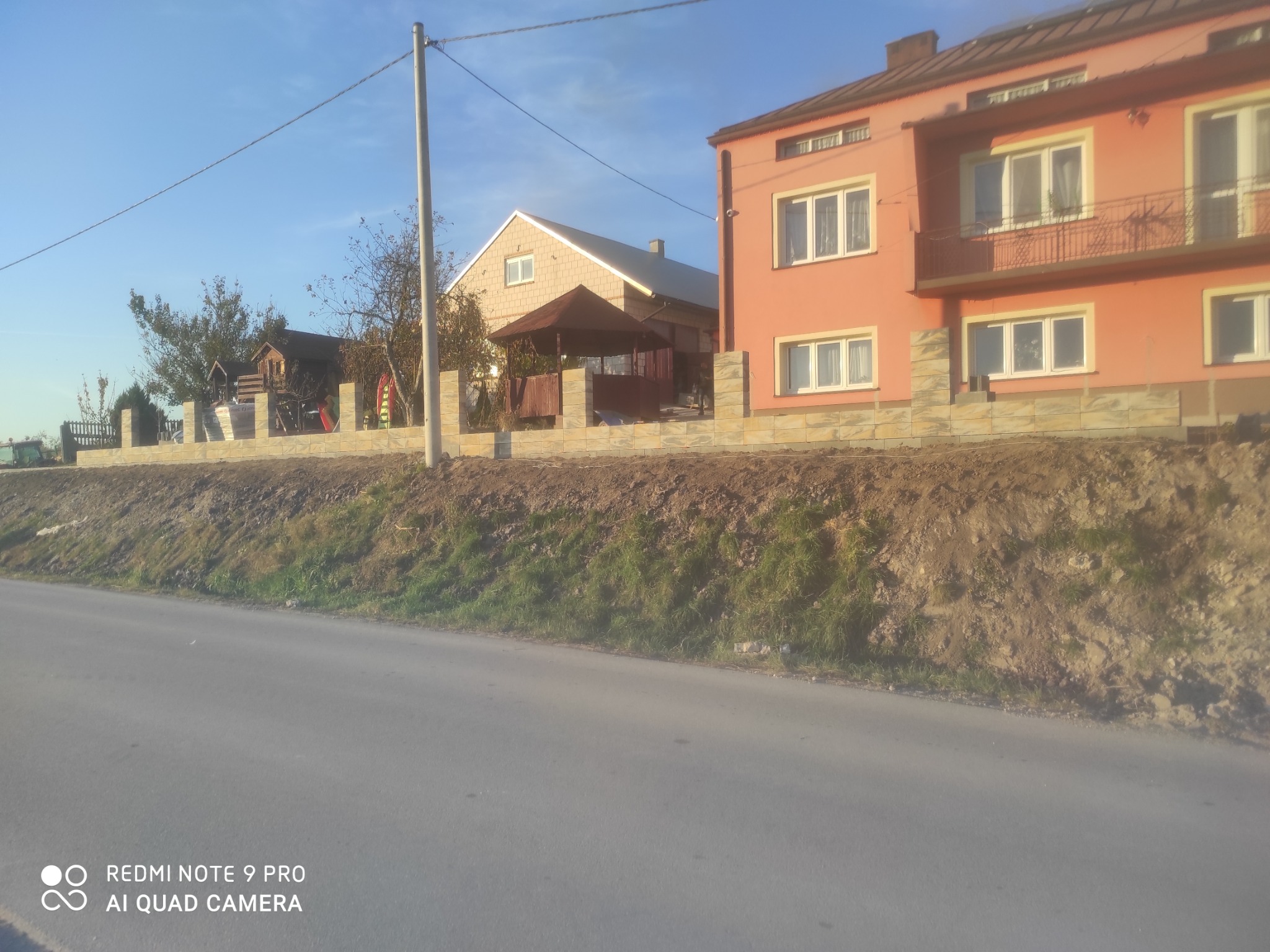 Niski murek z beżowych bloczków oddzielający posesję z domem w kolorze łososiowym oraz altaną od drogi, widok z poziomu jezdni.