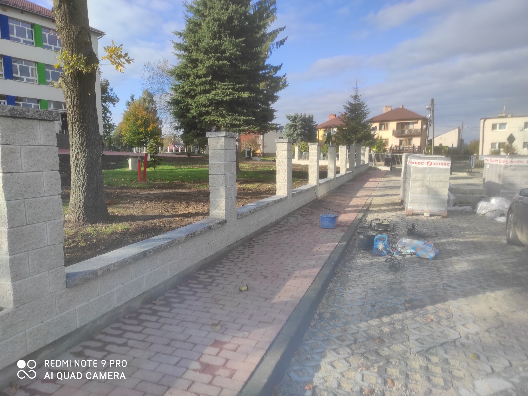 Wzniesiony mur z szarej kostki betonowej z kolumnami, oddzielający chodnik z czerwonej kostki brukowej od trawnika z drzewami, w tle budynek szkolny i domy jednorodzinne, palety z kostką brukową...