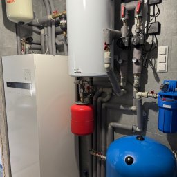 BWA Usługi Hydrauliczne - Złożona instalacja grzewcza w kotłowni: biały piec, zbiorniki (czerwony, niebieski, beżowy), rury w izolacji, zawory i sterowniki na tle szarej ściany z płytek.
