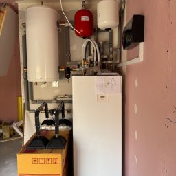 BWA Usługi Hydrauliczne - Biała pompa ciepła Mitsubishi, zbiornik buforowy i instalacja hydrauliczna na ścianie w surowym pomieszczeniu. Widoczny karton z pompą.