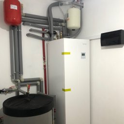 BWA Usługi Hydrauliczne - Biała pompa ciepła Rotenso Aquami z szarymi rurami i czerwonym zbiornikiem wyrównawczym w kotłowni. Widoczne zawory i manometry. Oświetlenie sufitowe.