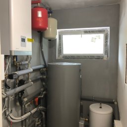 BWA Usługi Hydrauliczne - Kotłownia z białym piecem Rotenso, czerwonym i beżowym zbiornikiem IBO, szarym zasobnikiem i białym Galmet. Rury w izolacji. Okno Salamander w tle.