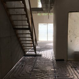 BWA Usługi Hydrauliczne - Montaż ogrzewania podłogowego w surowym wnętrzu. Widoczne rury na folii izolacyjnej, drewniane schody i betonowe ściany w trakcie budowy.