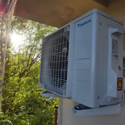 Zewnętrzna jednostka klimatyzacji Daikin zamontowana na elewacji budynku, widoczna siatka zabezpieczająca oraz zieleń drzew w tle, oświetlona promieniami słonecznymi.