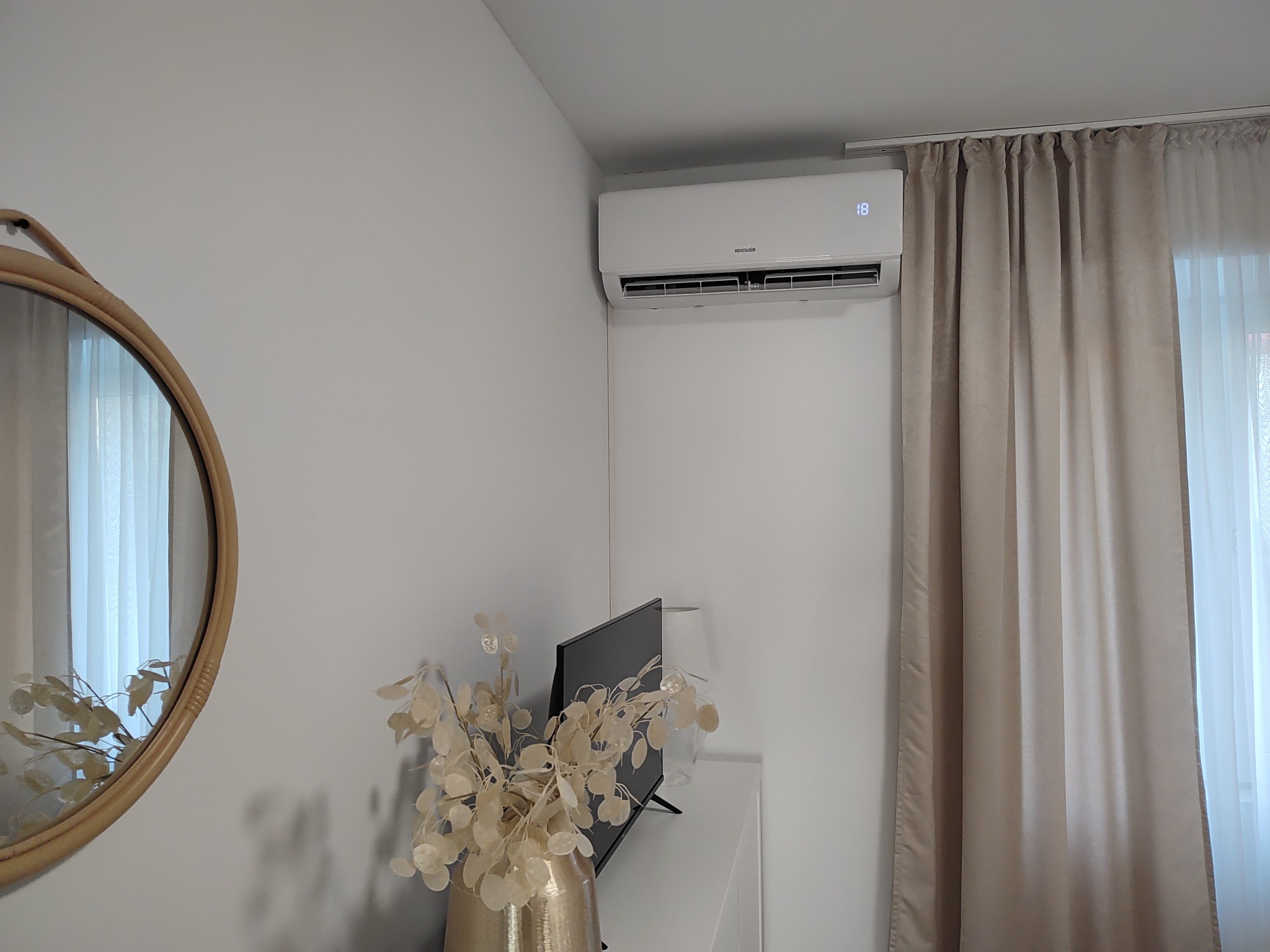 Biały klimatyzator ścienny marki Sinclair w nowoczesnym wnętrzu, z wyświetlaczem temperatury ustawionym na 18 stopni Celsjusza, zawieszony nad telewizorem i dekoracyjnym wazonem z suszonymi...