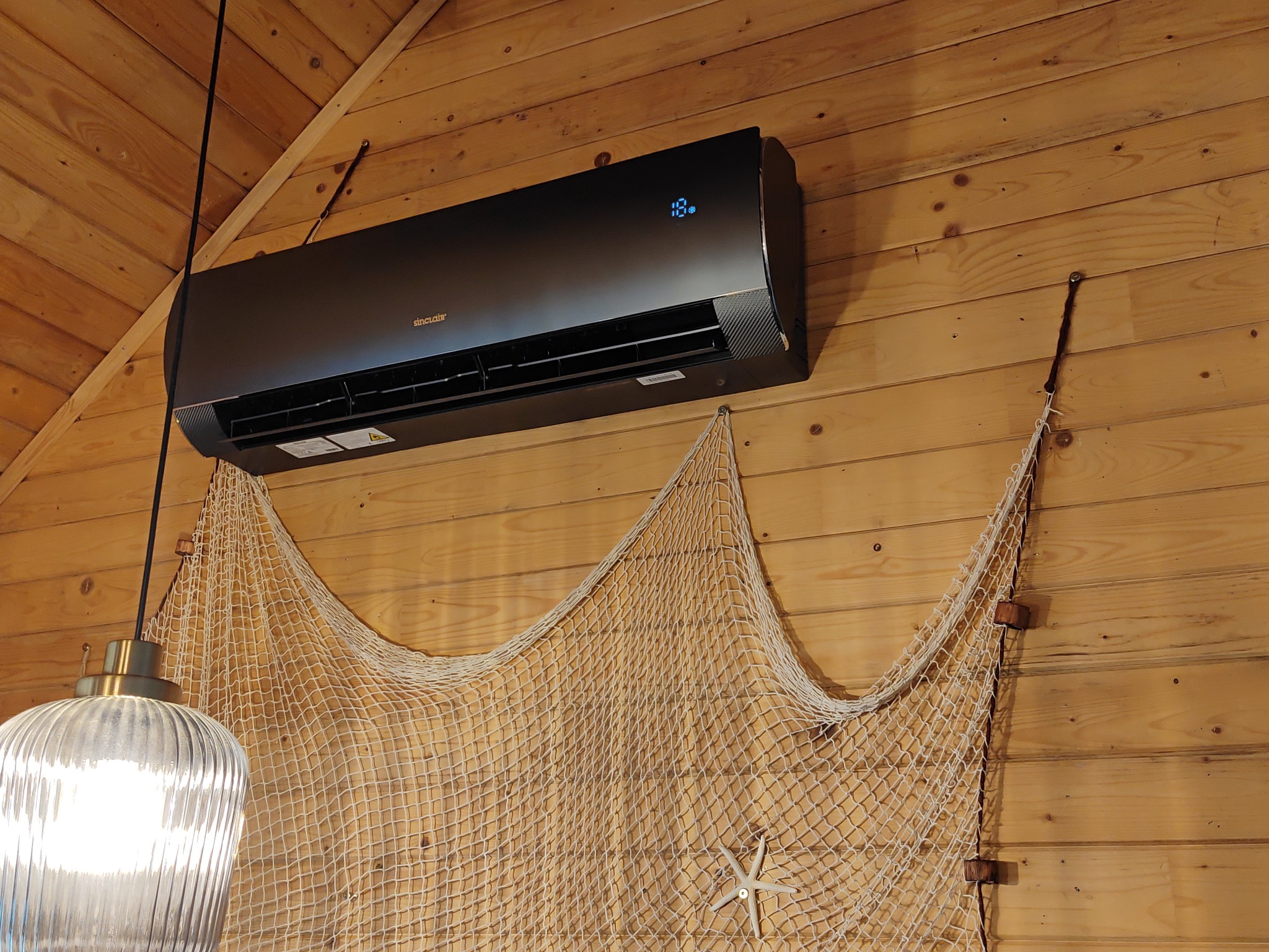 Czarna jednostka klimatyzacji Sinclair zamontowana na drewnianej ścianie, z dekoracyjną siecią rybacką i rozgwiazdą poniżej, oświetlona wiszącą lampą z prążkowanym kloszem.