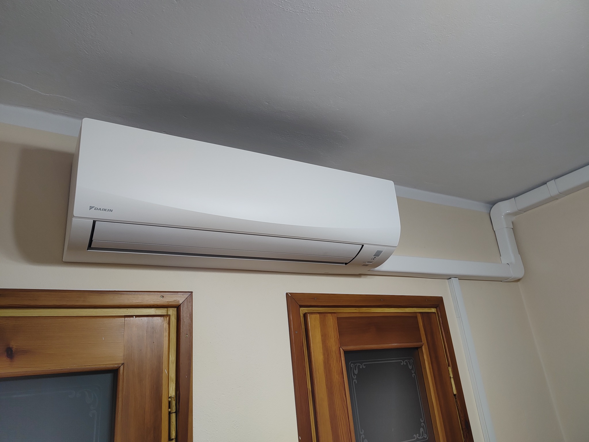 Biała jednostka klimatyzacji Daikin zamontowana na ścianie nad dwoma drewnianymi drzwiami z szybami, widoczne białe kable instalacyjne pod sufitem.