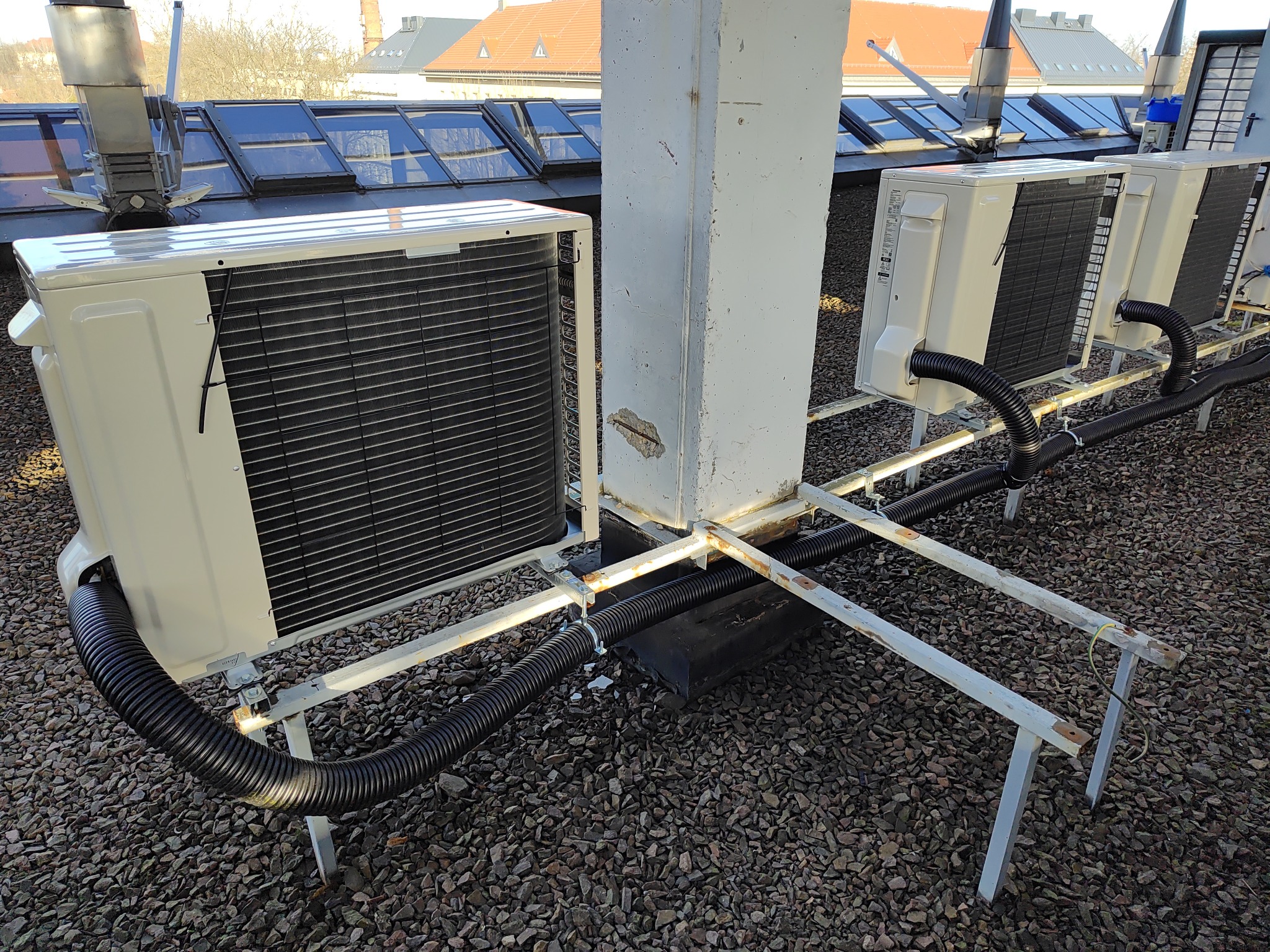 Zewnętrzne jednostki klimatyzacyjne zamontowane na metalowej konstrukcji na dachu budynku z widocznymi świetlikami i kominami w tle.