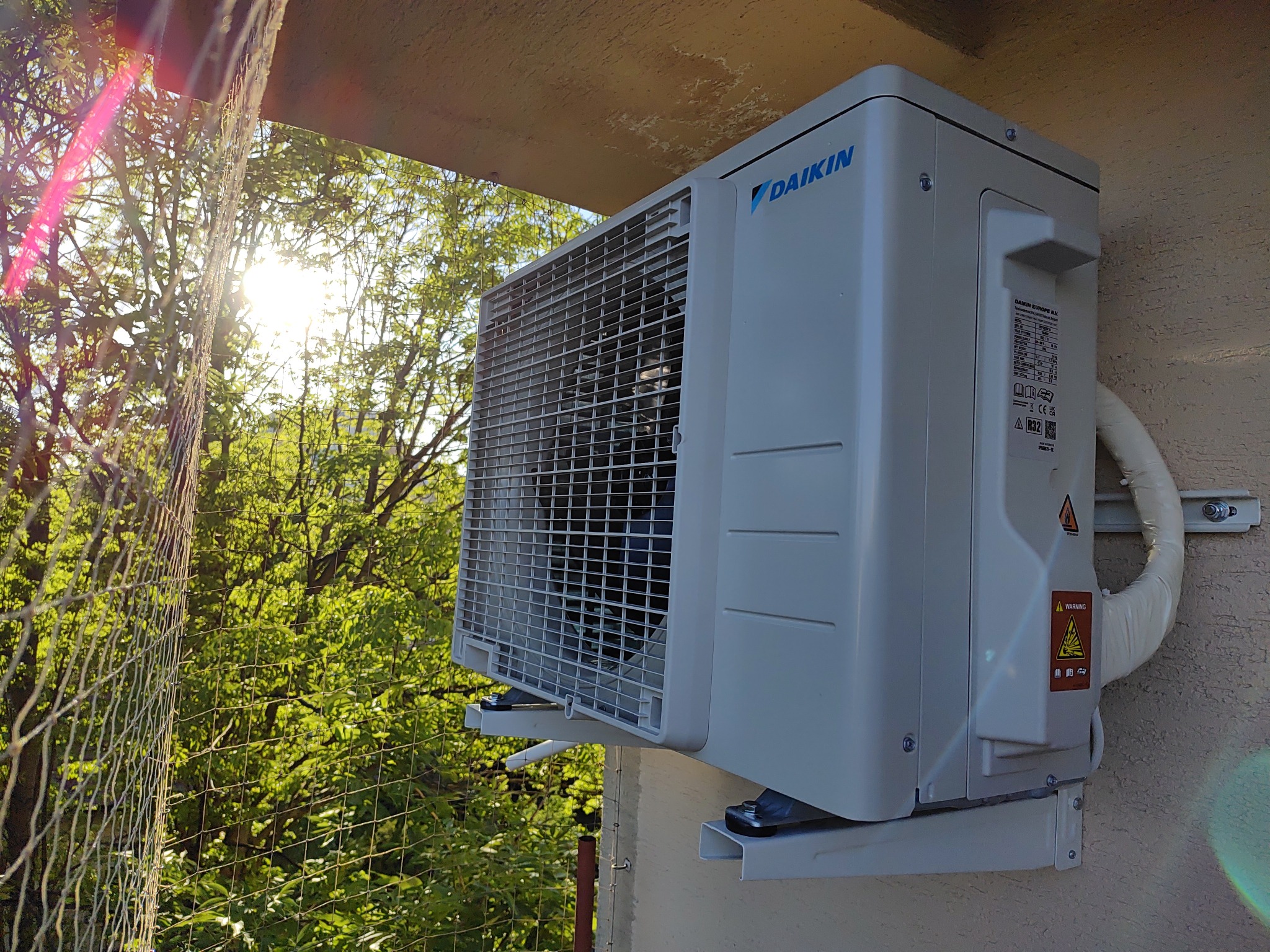 Zewnętrzna jednostka klimatyzacji Daikin zamontowana na elewacji budynku, widoczna siatka zabezpieczająca oraz zieleń drzew w tle, oświetlona promieniami słonecznymi.