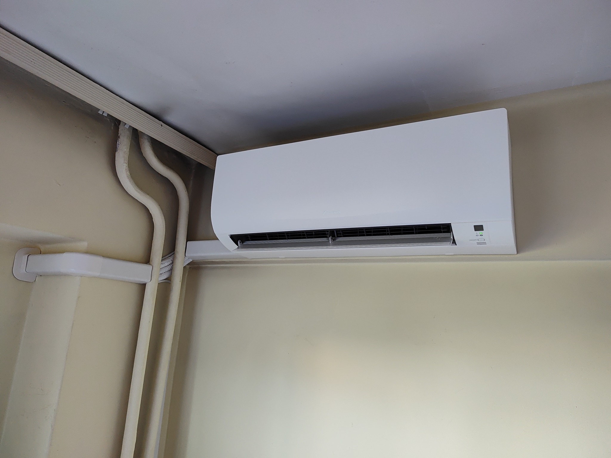 Biała jednostka klimatyzacyjna zamontowana na ścianie w pomieszczeniu, widoczne białe rury instalacyjne biegnące wzdłuż sufitu i ściany.