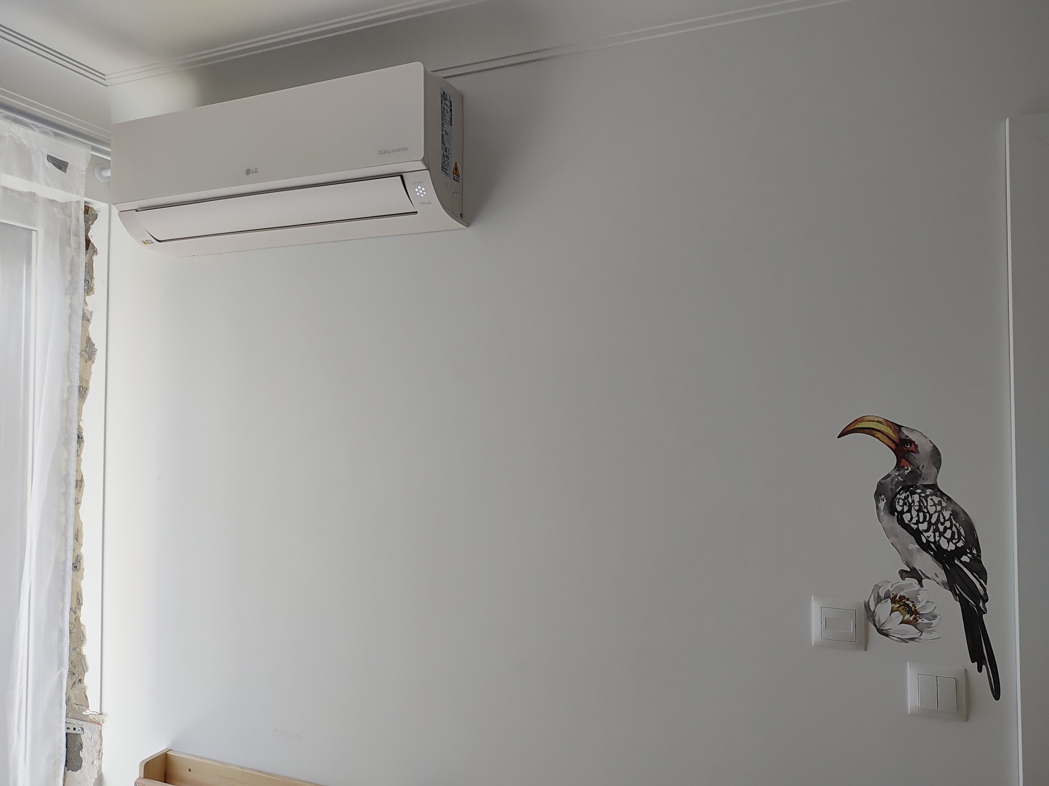 Biały klimatyzator LG zamontowany na ścianie pokoju z naklejką przedstawiającą ptaka.