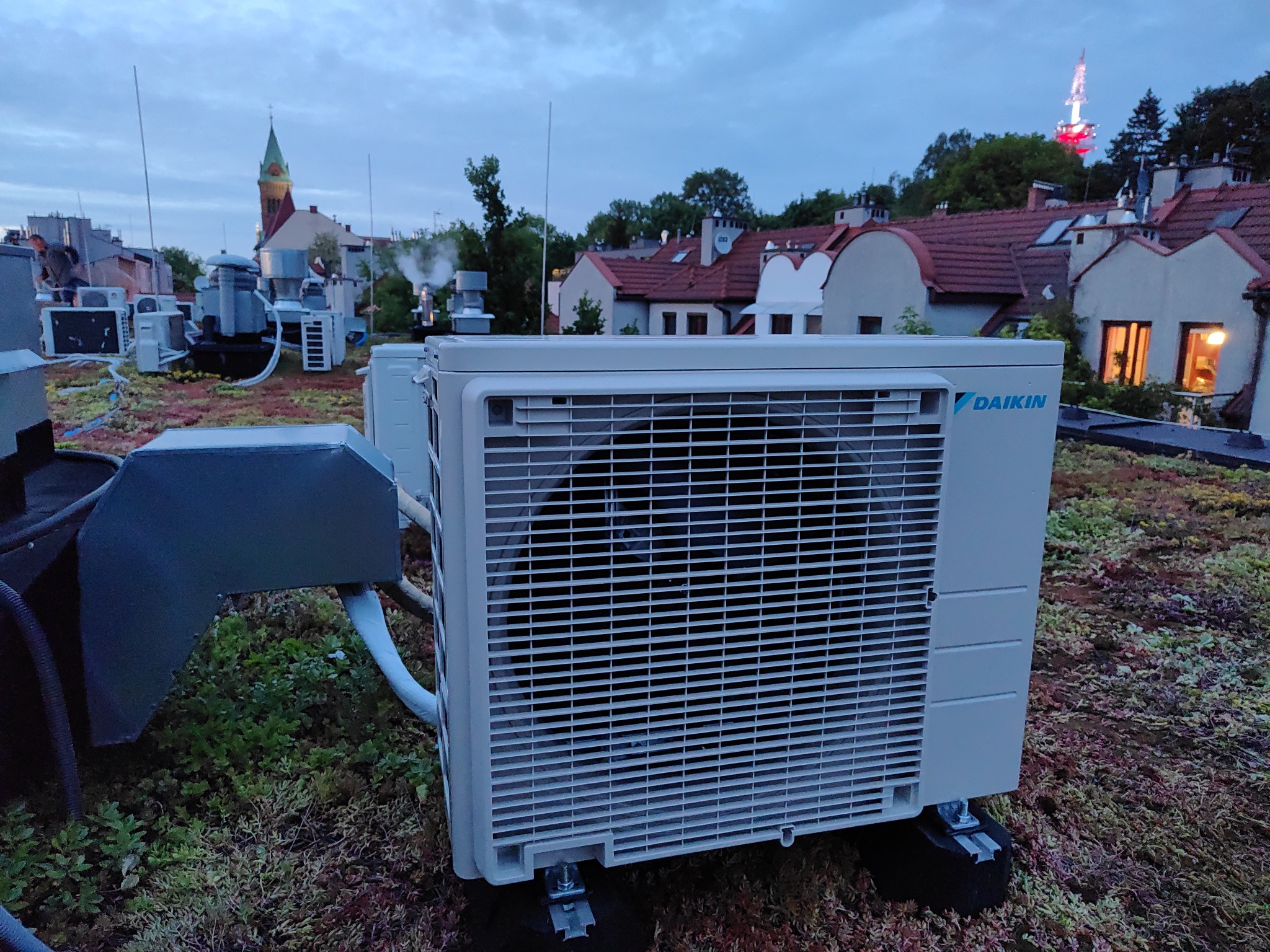 Jednostka zewnętrzna klimatyzacji Daikin na zielonym dachu budynku miejskiego, widok na kominy i wieżę telewizyjną w oddali.