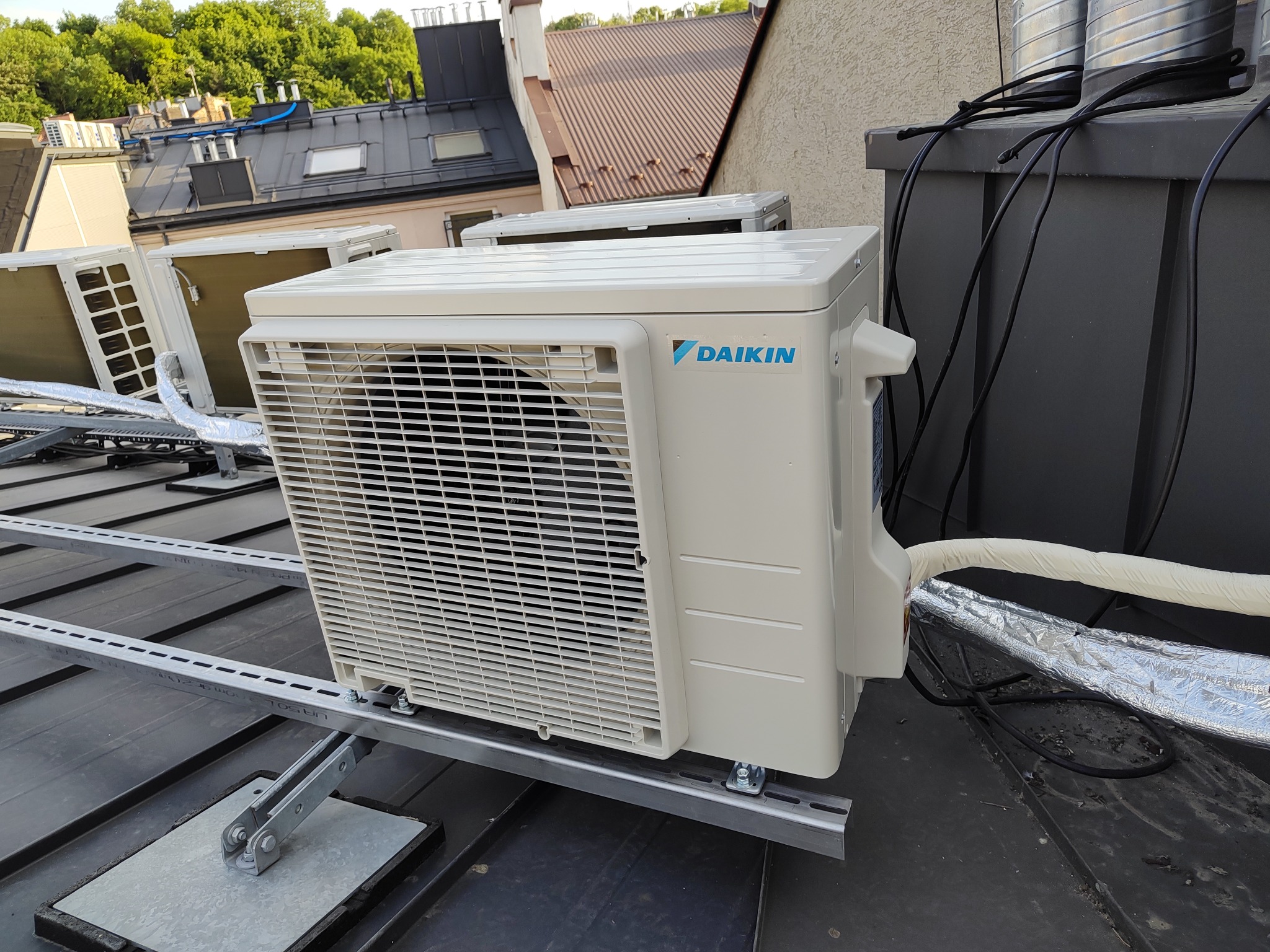 Zewnętrzna jednostka klimatyzacji Daikin zamontowana na dachu budynku z widocznymi elementami montażowymi i przewodami, na tle innych budynków i zieleni.