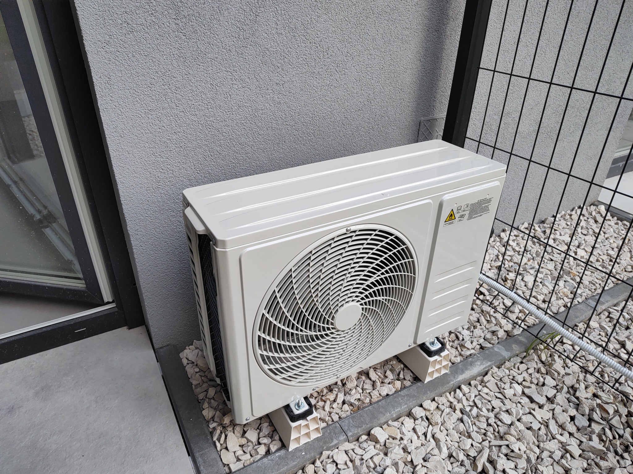 Zewnętrzna jednostka klimatyzacji zamontowana na białych wspornikach na tle szarej ściany i żwirowego podłoża z widoczną siatką ogrodzeniową.