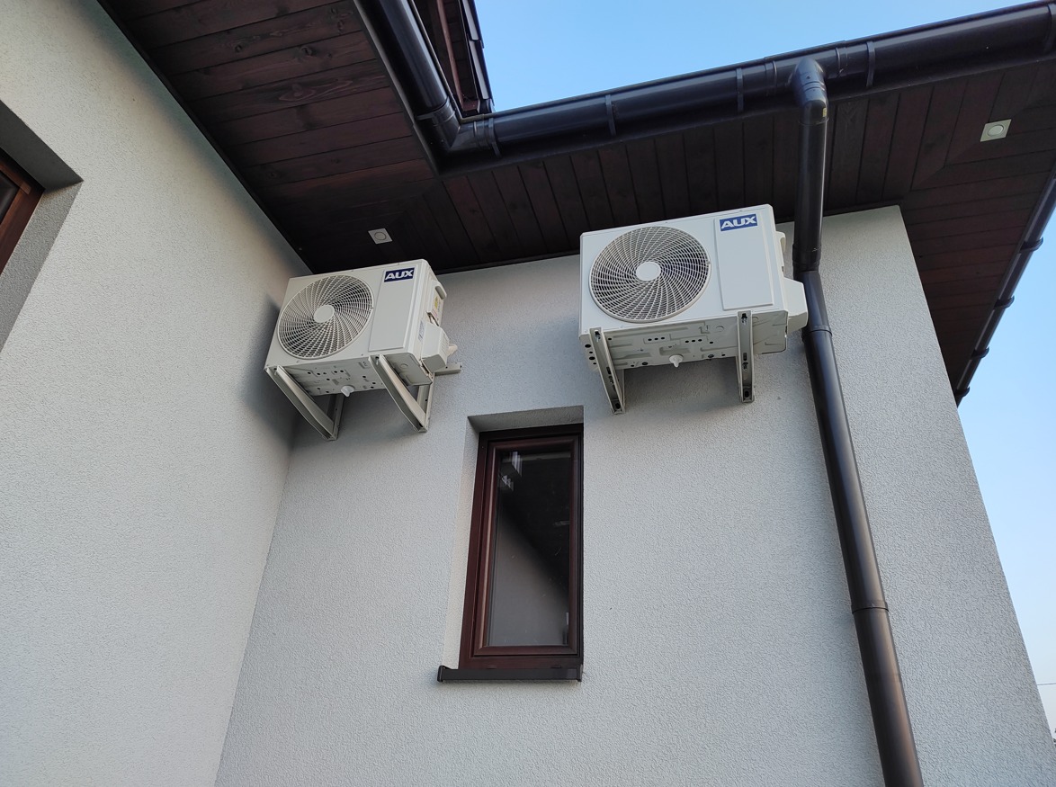 Dwa białe klimatyzatory marki AUX zamontowane na szarej, teksturowanej ścianie budynku obok okna i rynny dachowej.
