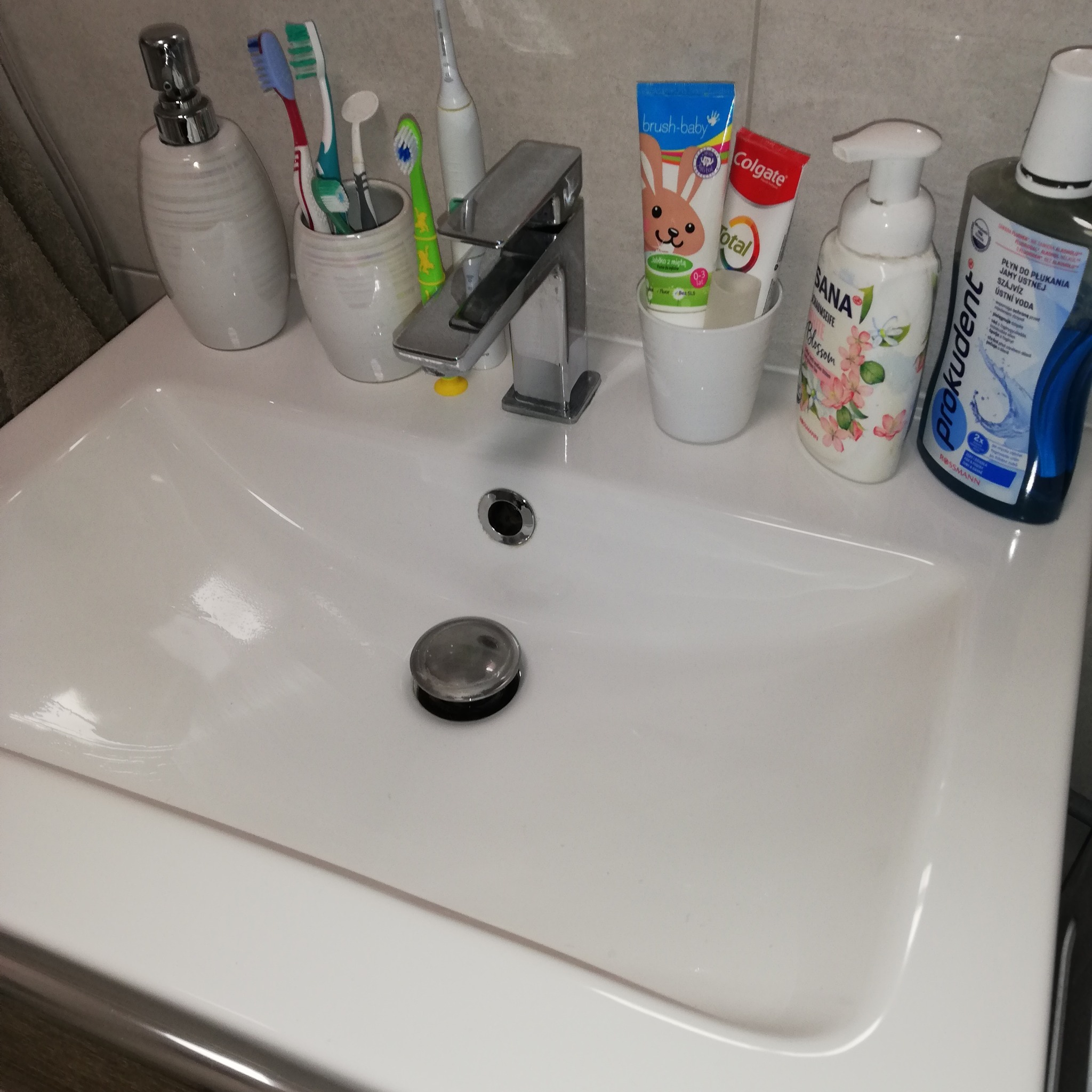 Biała umywalka z chromowaną baterią, obok której stoją pojemniki z pastami do zębów, szczoteczkami, mydłem w płynie i płynem do płukania jamy ustnej. Widoczne ślady po czyszczeniu.