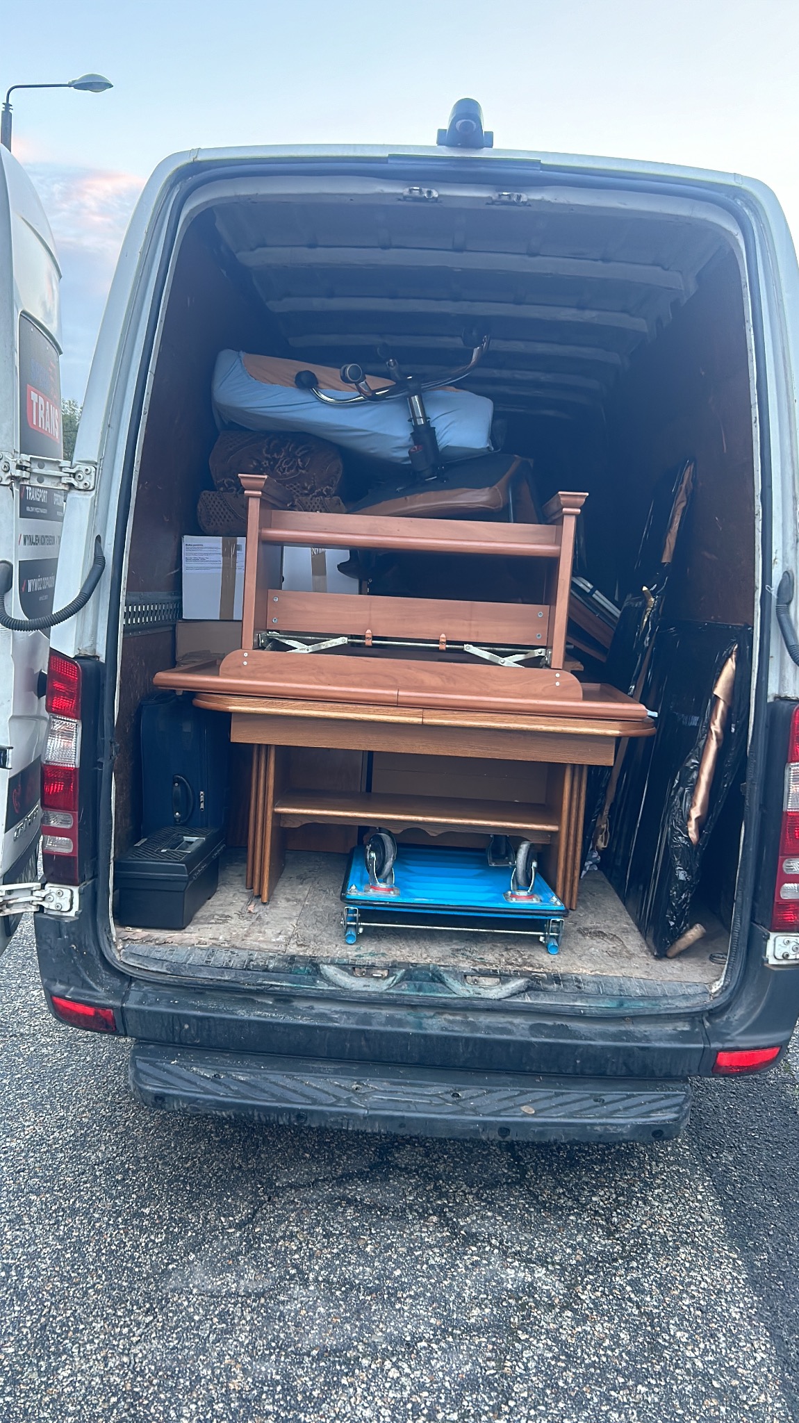 Wnętrze busa załadowane meblami, w tym rozłożonym drewnianym stołem, krzesłem biurowym i innymi przedmiotami przygotowanymi do transportu. Widoczny wózek transportowy pod stołem.