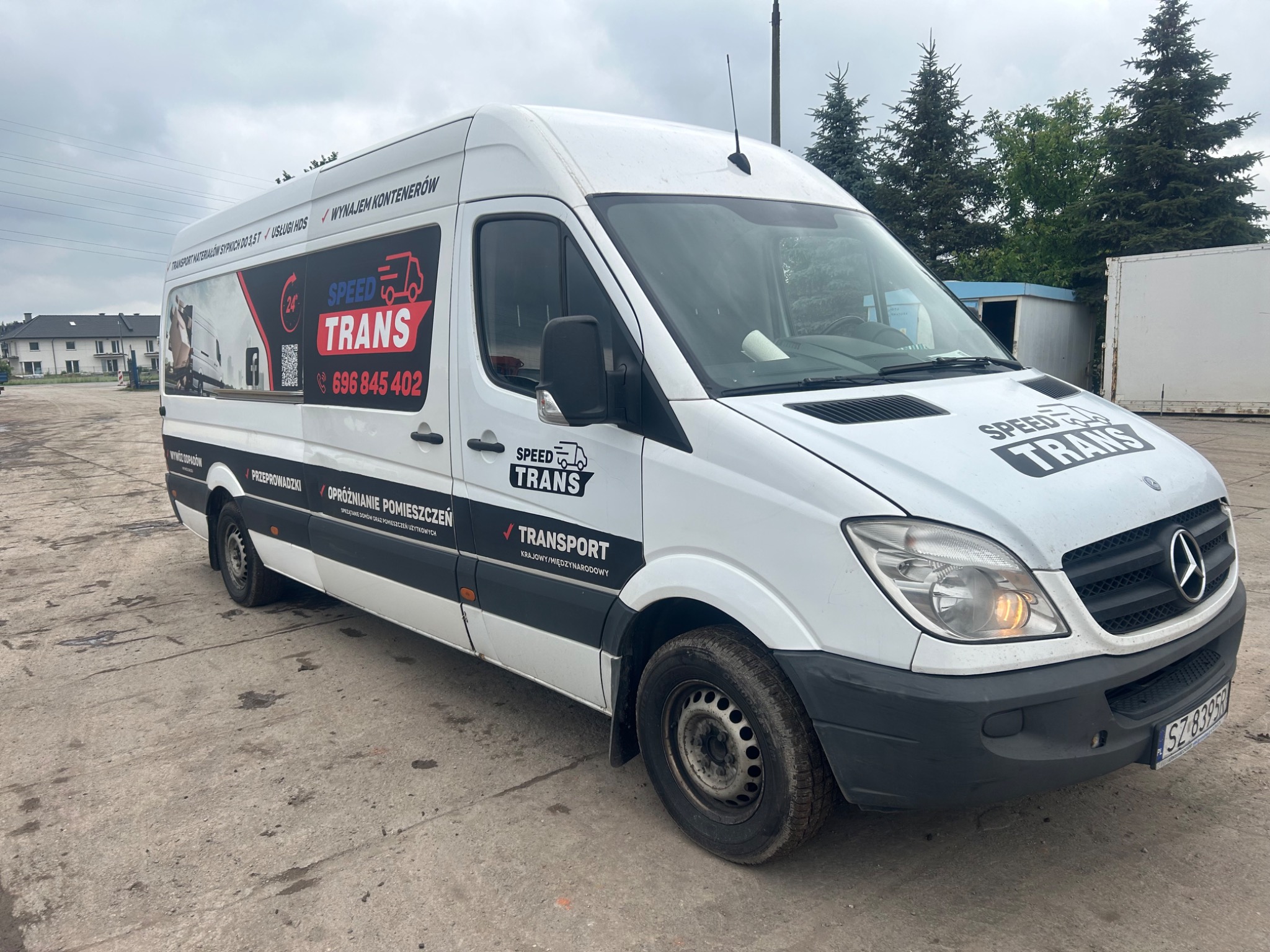 Biały bus firmy transportowej Speed Trans z widocznym logo i numerem telefonu, zaparkowany na betonowym placu. Na boku busa naklejki informujące o transporcie materiałów sypkich, wynajmie...