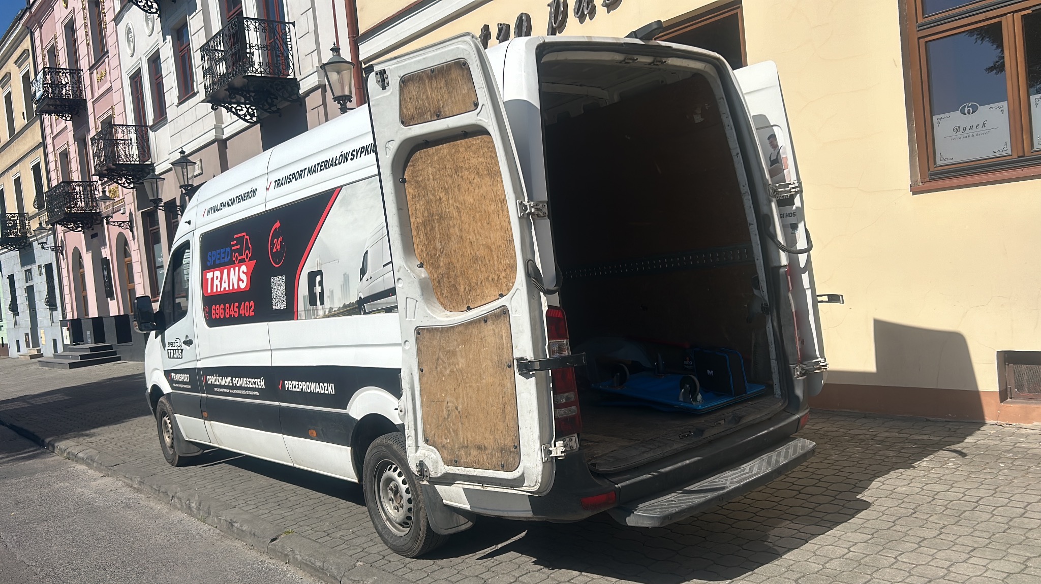 Biały samochód dostawczy z otwartymi tylnymi drzwiami na tle kamienic. Boczne drzwi samochodu oklejone reklamą firmy transportowej z numerem telefonu i informacją o transporcie, opróżnianiu...