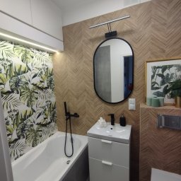 Chaykovskyy Group uslugi remontowe - Nowoczesna łazienka z wanną obudowaną szarą mozaiką, umywalką na szafce z dwoma szufladami, owalnym lustrem i płytkami imitującymi drewno ułożonymi w jodełkę na ścianie, z akcentem tropikalnych...