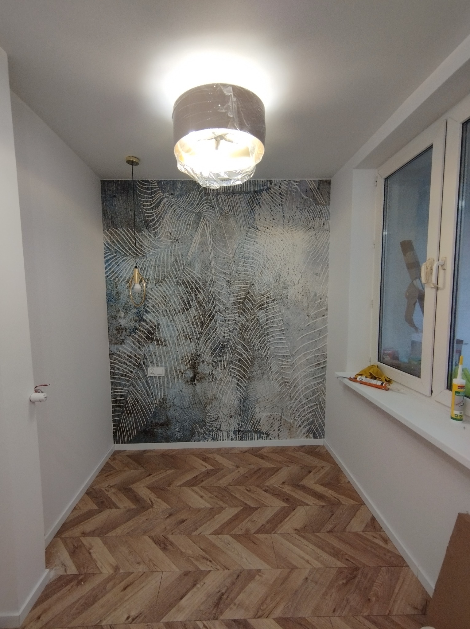 Pokój po remoncie: wzorzysta tapeta na ścianie, parkiet w jodełkę na podłodze, okno z parapetem, na którym leżą narzędzia i materiały budowlane, oraz lampa sufitowa w folii ochronnej.